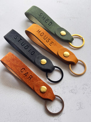 Handmade personalised leather keyrings by Parkin & Lewis, featuring embossed labels for car, house, studio, and shed keys, in orange, yellow, olive green, and navy leather with brass bronze hardware
