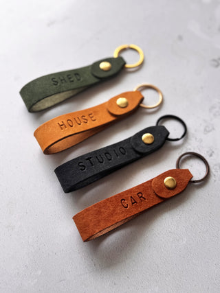 Handmade personalised leather keyrings by Parkin & Lewis, featuring embossed labels for car, house, studio, and shed keys, in orange, yellow, olive green, and navy leather with brass and bronze hardware