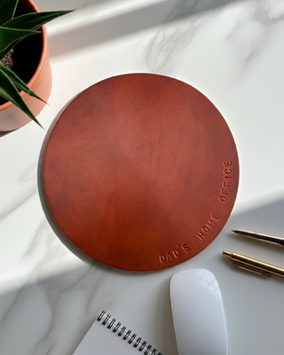 Round brown leather coaster with 'Dad's Home Office' engraving on a marble surface with a plant and pens.