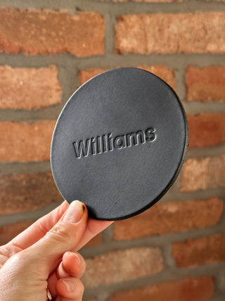 Black round lid with 'Williams' branding held against a brick wall.