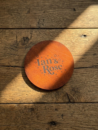 A round tan leather coaster with the words 'Ian & Rose' and small star illustrations, placed on a rustic wooden tabletop with diagonal sunlight and shadows.