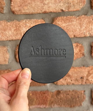 Black round coaster with 'Ashmore' branding held against a brick wall.