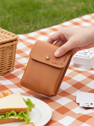 Tan leather card holder held by a hand on a picnic blanket with a sandwich and picnic basket.
