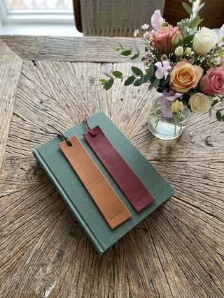 Two leather bookmark tags, one tan and one burgundy, attached to a green hardcover book and styled on a rustic wooden table next to a glass vase of flowers.