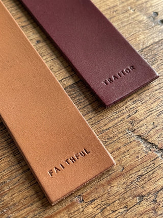 Two leather strap-like accessories laid on a rustic wooden surface: a tan one embossed with the word FAITHFUL and a burgundy one embossed with TRAITOR.