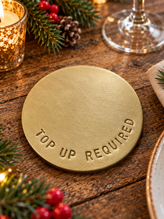 A round, brushed gold metal coaster engraved with the words 'TOP UP REQUIRED' sits on a rustic wooden table surrounded by holiday decorations like pine branches, red berries, a lit candle, and a wine glass base.