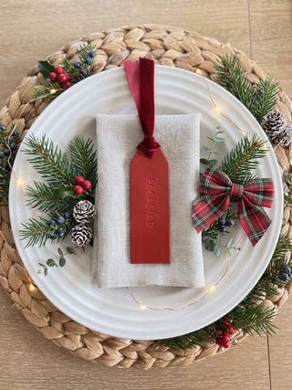 A festive holiday table setting featuring a white ceramic plate on a woven straw placemat, decorated with pine branches, berries, and fairy lights. In the center is a folded cream linen napkin with a red velvet ribbon and a matching red embossed gift tag reading 'FAGOLLIND'.
