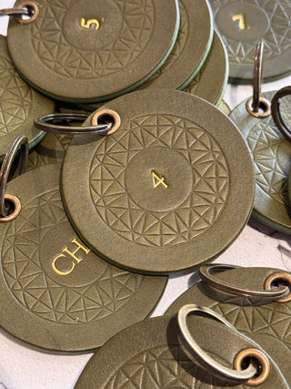 A close-up of multiple olive green leather key tags with embossed geometric patterns and gold-colored metal key rings, some showing gold numbers and letters.