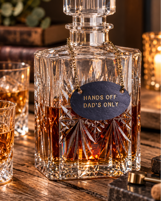A clear cut-crystal decanter with amber whiskey inside, styled on a wooden table with two matching crystal glasses. The decanter has a dark slate tag with gold lettering that reads 'HANDS OFF DAD'S ONLY'.