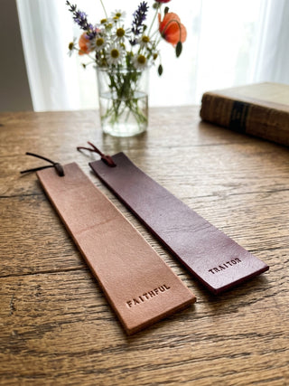 Two leather bookmark-like strips with tassels rest on a wooden table. One is light tan and embossed with the word FAITHFUL, and the other is darker reddish-brown with the word TRAITOR.