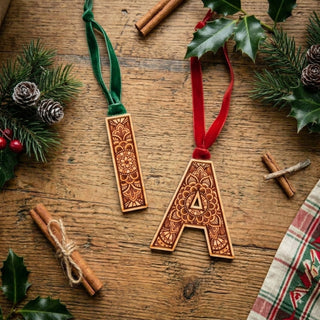 Two decorative wooden letter ornaments, one shaped like a rectangle and the other like a letter A, both with intricate laser-cut floral mandala patterns, hanging from velvet ribbons (green and red) on a rustic wooden table surrounded by Christmas greenery, pinecones, and cinnamon sticks.