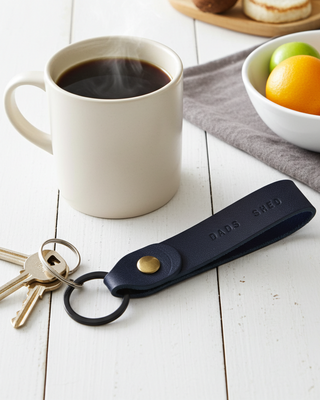 Navy blue keychain with keys on a white surface next to a mug of coffee and fruit bowl.