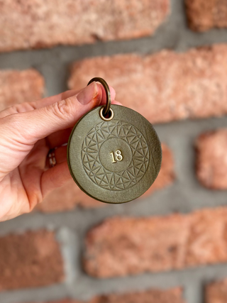 A hand holding a round olive green leather key tag with an embossed geometric pattern and the number 18 in the center, set against a blurred brick wall background.