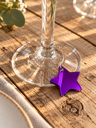 A metallic purple star-shaped wine glass charm resting on the base of a clear glass wine glass, set on a rustic wooden table with warm sunlight and bokeh background elements.