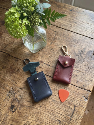 Two small leather music accessory pouches with metal clips are arranged on a rustic wooden table, alongside a small orange guitar pick and a glass vase with green flowers. One pouch is dark blue and the other is burgundy red.