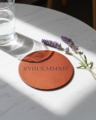 Tan Round leather coaster with engraved date on a marble surface with a glass of water and lavender.