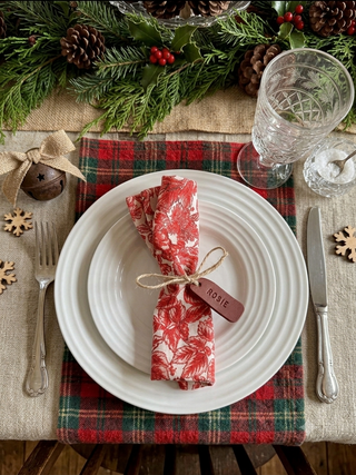 A festive holiday table setting featuring a red and white patterned napkin tied with twine and a small wooden name tag that reads 'ROSIE', placed on a white plate over a red and green plaid placemat. The setting includes silver cutlery, a clear glass, a small wooden ornament, pinecones, evergreen branches, and red berries on a burlap table runner.