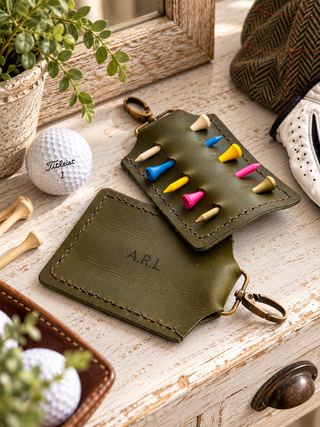 An olive green leather tee holder golf keychain organizer shown in a styled flat-lay setting with golf props, including a white Titleist golf ball, wooden tees, and a herringbone hat on a distressed white wooden surface.