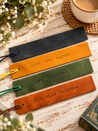 Four leather bookmark-style tags with colorful cords are arranged on a rustic wooden surface. The embossed text reads 'LEATHER ANNIVERSARY', 'LOVE YOU DADDY', 'DAY DREAMING', and 'OUR NEXT CHAPTER'.