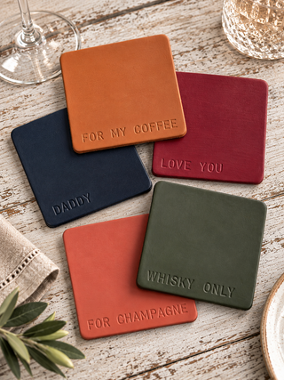 Five square leather coasters in orange, navy, burgundy, terracotta, and olive green, each embossed with a phrase like 'FOR MY COFFEE' or 'LOVE YOU', arranged on a rustic white-washed wooden surface with wine glass and plate props.