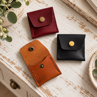 Three small leather card cases or coin purses in burgundy, tan, and black, arranged on a distressed white wooden surface with decorative plants and books around them.