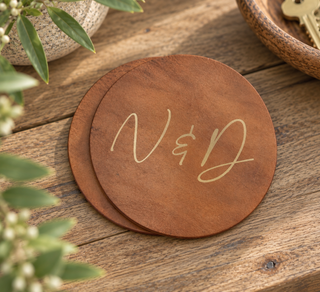 Two round wooden coasters stacked on a rustic wooden table, with the coasters engraved with the initials 'N & D' in a light cream script. The scene is styled with green leaves and a small ceramic pot in the foreground and background.