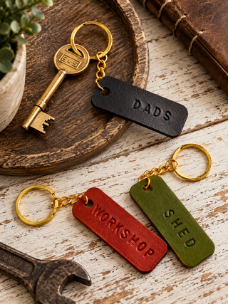 Three leather keychains with gold-tone keys and rings are arranged on a rustic white wood surface with a wooden tray, wrench, plant, and book as props. The keychain tags are black, red, and olive green, embossed with the words 'DADS', 'WORKSHOP', and 'SHED' respectively.