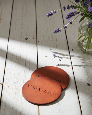 Two tan round leather coasters with engraved text on a wooden surface with lavender flowers.