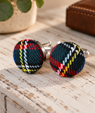 A pair of round cufflinks with colourful tartan plaid faces, resting on a distressed white wooden surface with a blurred potted plant and leather box in the background.