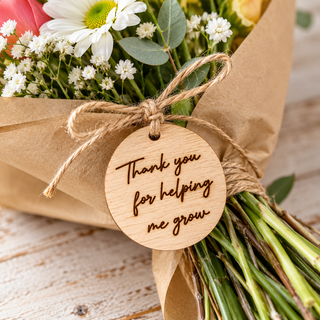 A close-up of a wrapped flower bouquet with brown kraft paper wrapping, twine, and a round wooden tag that reads 'Thank you for helping me grow.' The bouquet includes white daisies, small white filler flowers, green leaves, and pink and yellow flowers on a rustic white wood surface.