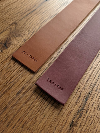 Two leather belt straps laid diagonally on a wooden surface, one tan with the word FAITHFUL embossed and one muted burgundy with the word TRAITOR embossed.