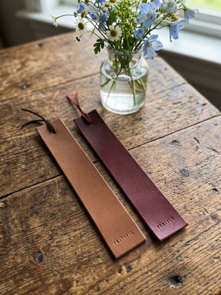 Two slim leather bookmarks or name tags with tassels lie on a rustic wooden table. One is light tan and the other is dark burgundy, both with small leather loops and the names 'FAITHU' and 'TRAITOR' embossed near the bottom.FUL