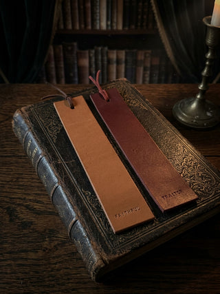 Two leather bookmark bookmarks with tassels resting on an antique book on a wooden table, with a candlestick and bookshelves in the background. One bookmark is tan and reads 'FAITHFUL', and the other is reddish-brown and reads 'TRAITOR'.