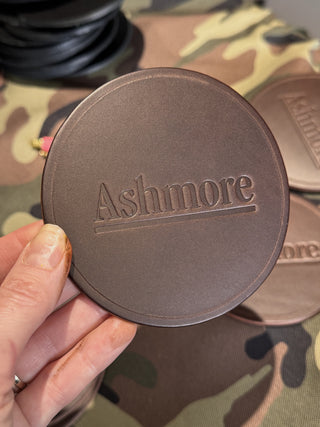 Round brown object with 'Ashimore' branding held by a hand on a camouflage-patterned surface.