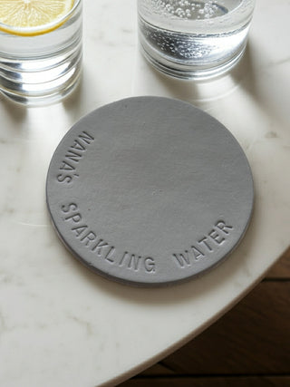 Round gray coaster with 'Nana's Sparkling Water' text on a marble surface with glasses of water.