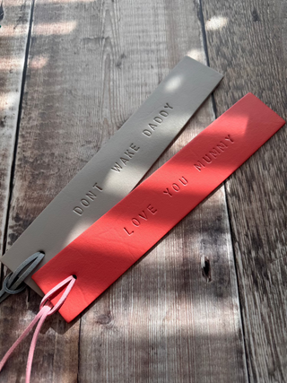 Two handcrafted leather bookmarks in putty and coral each embossed with sentimental messages: 'DONT WAKE DADDY' and 'LOVE YOU MUMMY,' ' Each bookmark has a matching leather tassel and is displayed on a rustic wooden surface with soft natural lighting