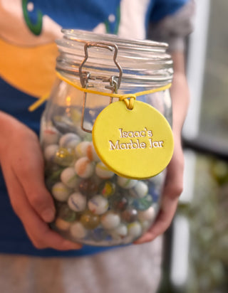 A glass jar wrapped with a hand-painted yellow label that reads "Isaac's Marble Jar" in neat, silver text. The label adds a personal and cheerful touch to the jar, which is likely used for collecting rewards or marbles