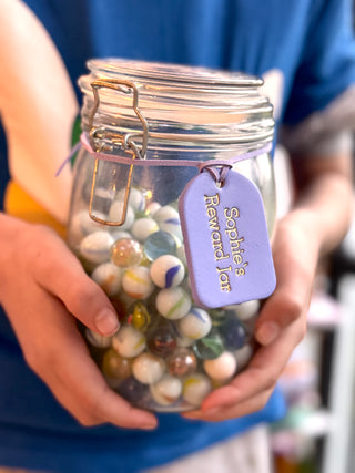 A marble jar wrapped with a hand-painted lavender purple label that reads "Sophie's Reward Jar" in neat, handwritten-style lettering. The label adds a personal and cheerful touch to the jar, which is likely used for collecting rewards or incentives