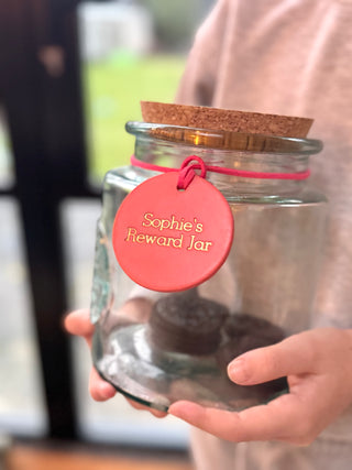 A glass jar wrapped with a hand-painted coral pink label that reads "Sophie's Reward Jar" in neat, handwritten-style lettering. The label adds a personal and cheerful touch to the jar, which is likely used for collecting rewards or incentives, or a label for a biscuit tin.
